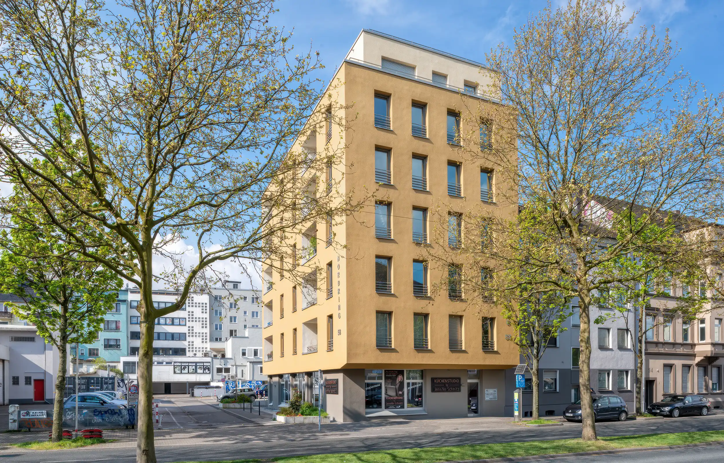 „Modernes mehrstöckiges Wohngebäude mit gelber Fassade und großen Fenstern, umgeben von Bäumen und anderen Gebäuden in einer städtischen Umgebung.